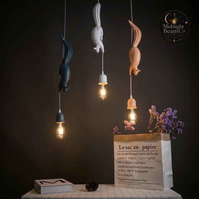 May include: Three squirrel-shaped pendant lights in navy, white, and peach. Each light has a decorative cap and a vintage-style bulb. The lights hang from white cords against a dark background. A paper bag with French text and purple flowers are in the foreground.
