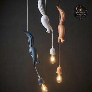 A warm pink and beige resin squirrel pendant lamp hanging from a matching cord with an acorn-shaped lamp cap and warm Edison bulb, showing the playful diving squirrel sculpture from a side angle against a dark backdrop.