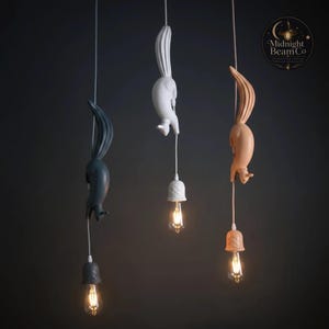 Op de afbeelding: Drie hanglampen met eekhoornvormige armaturen in grijs, wit en perzik. Elke eekhoorn hangt aan een snoer en houdt een klokvormige lampenkap met een vintage Edison-lamp. De lampen hangen tegen een donkere achtergrond.