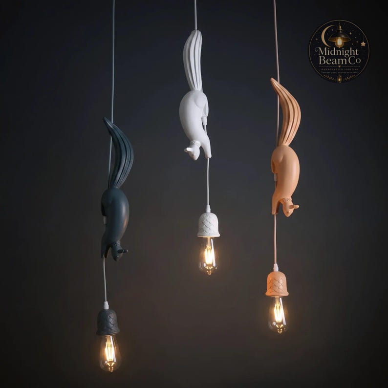 Three resin squirrel pendant lamps in matte white, slate blue, and warm pink hanging from the ceiling at varying heights, each squirrel diving headfirst with paws extended downward holding a glowing Edison bulb below.