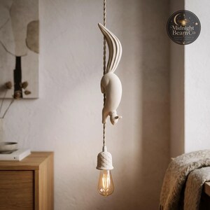 May include: A cream-colored squirrel pendant light fixture. The squirrel is sculpted in a climbing pose on a twisted cord, with a textured lampshade and a warm-toned bulb. The fixture is suspended in front of a neutral-toned wall. The logo for Midnight Beam Co is in the background.