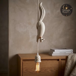 A slate blue resin squirrel pendant lamp suspended from a matching blue cord, showing the squirrel figure mid-dive with detailed fur texture, paws reaching toward an acorn lamp cap and warm glowing Edison bulb below.