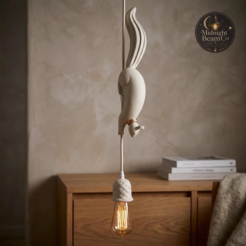 A slate blue resin squirrel pendant lamp suspended from a matching blue cord, showing the squirrel figure mid-dive with detailed fur texture, paws reaching toward an acorn lamp cap and warm glowing Edison bulb below.