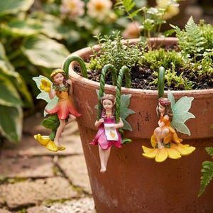 May include: Three miniature fairy figurines in a garden setting. Each fairy is dressed in a colorful dress and has delicate wings. They are perched on a flower pot, with one on a swing.