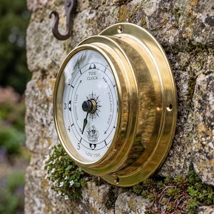 May include: A circular tide clock with a polished brass casing and a white face. The dial features black markings, a central compass rose, a small ship illustration, and text reading TIDE CLOCK, HIGH TIDE, LOW TIDE, RISING TIDE, and FALLING TIDE.