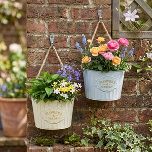 Könnte beinhalten: Zwei hängende Metalleimer mit Seilgriffen, einer cremefarben und einer hellblau, jeweils mit einem dekorativen Schild mit der Aufschrift "FLOWERS & GARDEN". Die Eimer sind mit bunten Blumen gefüllt, darunter rosa, orange und blaue Blüten.