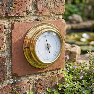 May include: A round brass hygrometer with a white face, black numerical markings from 0 to 100, and a central compass-style needle. The device features the word Hygrometer printed in black script at the bottom and is mounted on a brick surface.