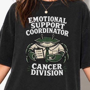 May include: A black short-sleeve t-shirt featuring a graphic of a green crab wearing a white shirt and tie, holding a clipboard. The text reads EMOTIONAL SUPPORT COORDINATOR CANCER DIVISION with the clipboard listing LISTEN, VALIDATE, SUPPORT.