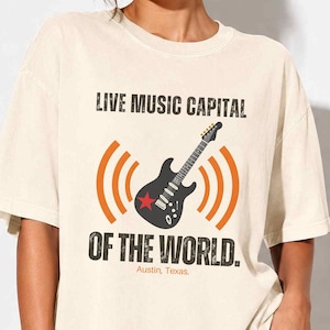May include: Ivory-colored t-shirt with the text "LIVE MUSIC CAPITAL OF THE WORLD" and a graphic of a black guitar with orange sound waves and a red star. The text "Austin, Texas" is below the graphic.