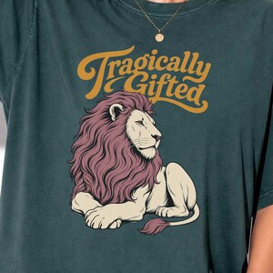 May include: A dark teal short-sleeve t-shirt featuring a graphic of a lion with a dark pink mane. Above the lion, the text "Tragically Gifted" is printed in a mustard yellow script font. The shirt has a crew neckline and a relaxed, loose fit.
