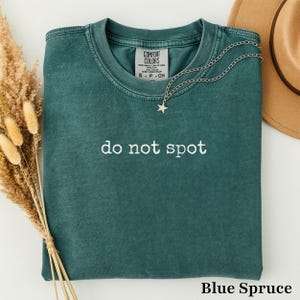 Puede incluir: Una camiseta de cuello redondo en color verde azulado oscuro con acabado lavado vintage. El pecho presenta el texto bordado en blanco 'do not spot'. Un collar de cadena plateada con un pequeño colgante de estrella descansa en el cuello y se ve un sombrero beige.