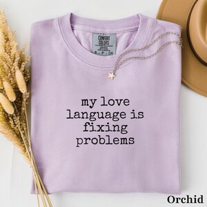 Pode incluir: Uma t-shirt de gola redonda em violeta claro com o texto preto "my love language is fixing problems" impresso na frente. A peça apresenta uma etiqueta da marca Comfort Colors no interior da gola e é acompanhada por um colar de estrela prateado.
