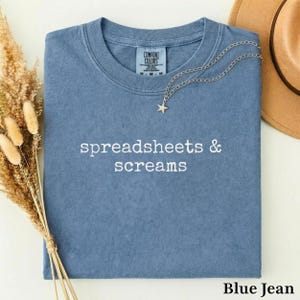 Può includere: Una t-shirt color blu jeans con scollo rotondo e la scritta "spreadsheets & screams" ricamata in bianco sul petto. La maglietta presenta un'etichetta del marchio Comfort Colors all'interno del collo e una collana con ciondolo a stella.