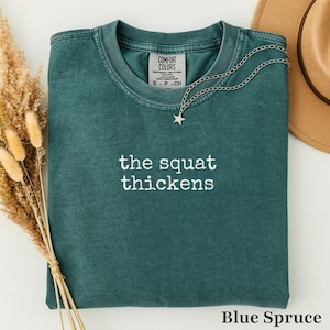 May include: A dark teal crewneck t-shirt with a vintage wash, featuring the white embroidered text "the squat thickens" centered on the chest. The shirt includes a Comfort Colors brand label at the inner neckline and a silver star pendant necklace.
