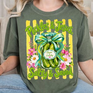 May include: An olive green t-shirt featuring a graphic of a glass jar filled with pickles, tied with a teal ribbon. The text reads Pickles & Jesus, Salty & Saved, and Chosen & Crunchy. The design includes yellow vertical stripes and pink and white flowers.