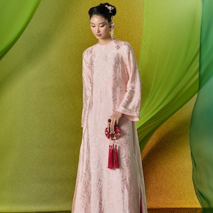 May include: A pale pink, long-sleeved dress with a floral design. The dress has a high neckline and a long, flowing skirt. The person is holding a red and gold decorative item with tassels. The background features green and yellow fabric.