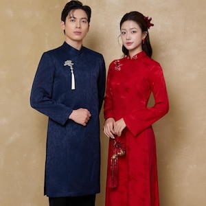 May include: A couple in traditional Asian attire. The person on the left wears a navy blue, long-sleeved tunic with a floral pattern, paired with black pants. The person on the right is in a red, long-sleeved dress with matching pants. Both outfits have mandarin collars.
