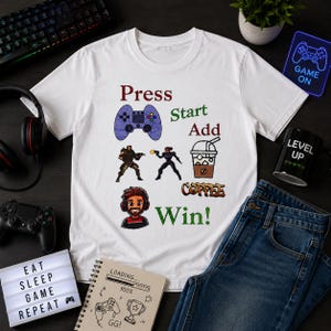 Könnte beinhalten: Weißes T-Shirt mit Gaming-Motiv. Das Shirt zeigt eine lila Gamecontroller-Grafik, Actionfiguren, eine Kaffeetasse und die Worte "Press Start Add Coffee Win!". Eine Jeans ist neben dem Shirt.