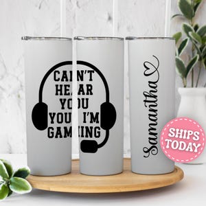 May include: A white cylindrical tumbler with a black graphic of gaming headphones and the text CAN'T HEAR YOU I'M GAMING. The side features the name Samantha in black script with a heart icon above it. The tumbler includes a clear plastic straw.