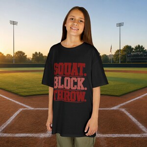 May include: A black short-sleeved t-shirt featuring the red text SQUAT. BLOCK. THROW. printed in a bold, distressed font on the front. The shirt has a crew neckline and a relaxed fit, providing a simple and functional design for athletic wear.