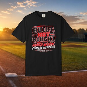 Puede incluir: Una camiseta negra de manga corta con el texto en rojo y blanco BUILT, NOT BOUGHT HARD WORK CHANGES EVERYTHING. Debajo del texto, se lee VANCOUVER RAVENS BASEBALL EST 2022. El diseño incluye detalles de pinceladas rojas.