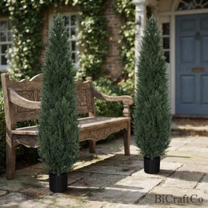 May include: Two tall, slender, conical artificial evergreen trees with dense, dark green needle-like foliage. Each tree is potted in a simple, cylindrical black container. The trees are positioned upright on a stone patio surface.