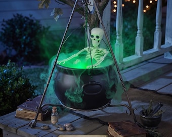May include: A black cauldron suspended from a metal tripod, featuring a skeleton figure inside with glowing green light and mist. A label at the base reads Witch Cauldron & Tripod - 52" Tall. The setup includes two leather-bound books and a small bottle.
