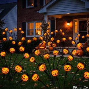 May include: A collection of illuminated orange jack-o'-lantern garden stakes with carved faces arranged in a grassy yard. The glowing pumpkin lights are positioned in front of a brick house with a porch light and a large carved pumpkin on the steps.