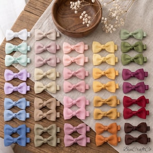 May include: A collection of small hair bows in various solid colors, including white, mint, lavender, blue, beige, pink, yellow, olive, burgundy, and brown. Each bow features a ribbed texture and is attached to a metal alligator hair clip.