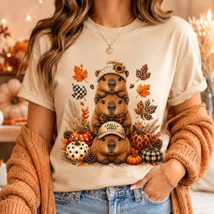 May include: A beige t-shirt featuring a graphic of three stacked capybaras. The top capybara wears a beanie with a sunflower, the middle wears a leopard print bow tie, and the bottom wears a cap with the text FALL VIBES. Surrounded by pumpkins and leaves.
