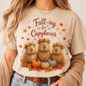 May include: A beige short-sleeved t-shirt featuring a graphic of three brown capybaras wearing floral crowns, surrounded by autumn leaves and pumpkins. The text "Fall-ing for Capybaras" is printed in brown script above the capybaras.