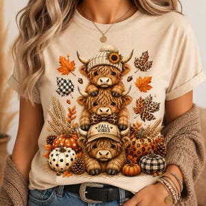 May include: A beige t-shirt featuring a graphic of three stacked Highland cows. The top cow wears a beanie with a sunflower, the middle wears a leopard print bow tie, and the bottom wears a cap with the text FALL VIBES. Surrounded by pumpkins, wheat, and autumn leaves.