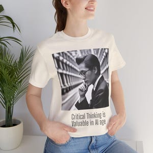 May include: A cream-coloured short-sleeve t-shirt featuring a black and white graphic of a person in a suit and glasses with their hand near their chin. Below the graphic, the text reads: Critical Thinking is Valuable in AI age.