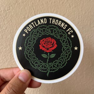 Puede incluir: Una pegatina redonda con el texto "PORTLAND THORNS FC" en blanco, arqueado en la parte superior. Presenta una rosa roja en el centro, rodeada por un dise&ntilde;o de vid espinosa verde sobre un fondo negro. Dos estrellas doradas flanquean el texto.
