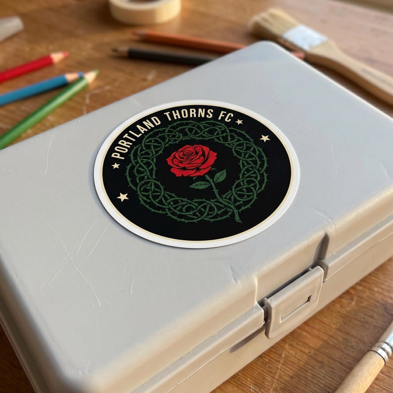 Puede incluir: Una pegatina redonda con el texto "PORTLAND THORNS FC" en el borde. La pegatina tiene un fondo negro con una corona de espinas verdes que rodea una rosa roja. Dos estrellas blancas flanquean el texto.