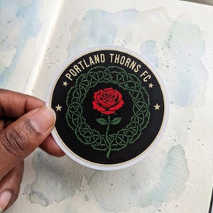 Puede incluir: Adhesivo redondo con la inscripci&oacute;n "PORTLAND THORNS FC" en blanco. El dise&ntilde;o presenta una rosa roja en el centro, rodeada por una vid espinosa verde sobre un fondo negro. Dos estrellas flanquean el texto.
