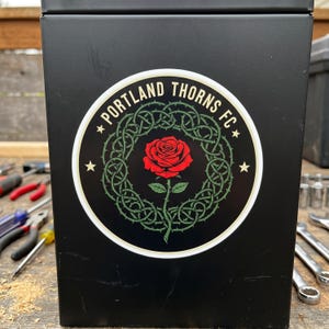 Puede incluir: Caja de herramientas de metal negro con un emblema circular. El emblema muestra el texto "PORTLAND THORNS FC", una rosa roja en el centro y un dise&ntilde;o de vid espinosa verde. Dos estrellas flanquean el texto.