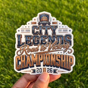 May include: A die-cut sticker featuring a shield emblem with a basketball and the text CITY LEGENDS, Road to Victory, CHAMPIONSHIP, and EST. 2026. The design uses a color palette of navy blue, metallic bronze, and white with star accents.