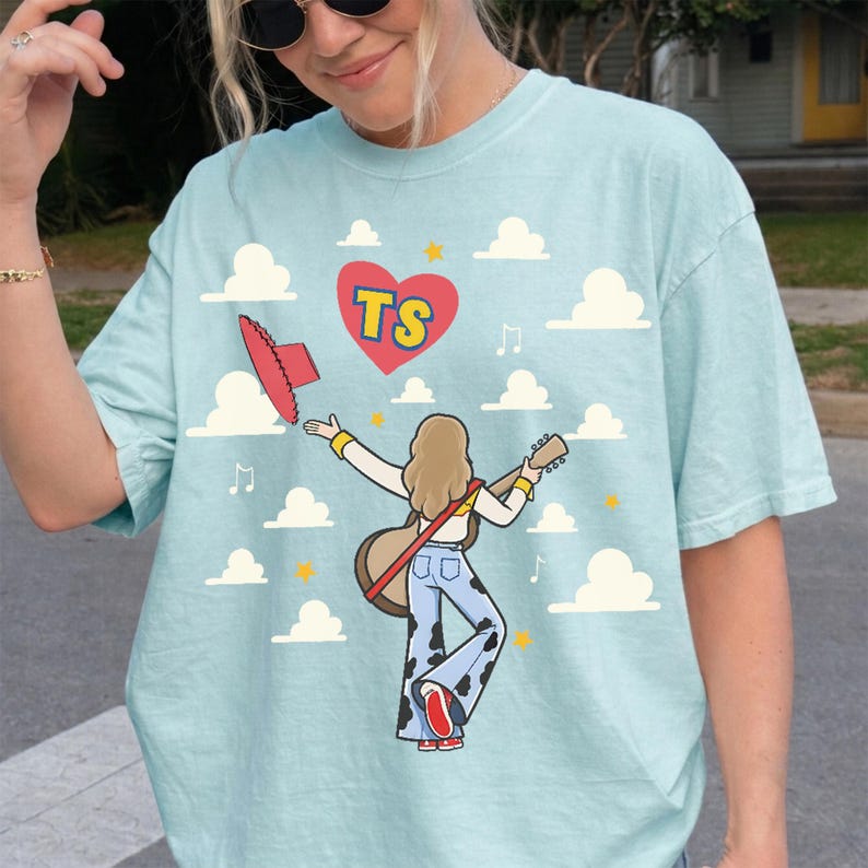 May include: A light blue t-shirt featuring a graphic of a person with long hair playing an acoustic guitar, wearing cow-print bell-bottom pants and a red hat. The design includes a red heart with the letters TS, white clouds, and yellow stars.