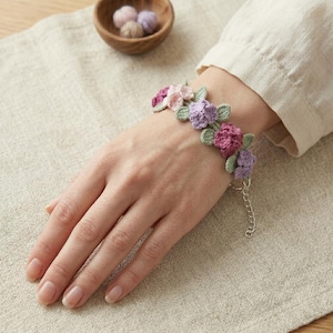 May include: A bracelet featuring a series of small, crocheted flowers in shades of pink, purple, and lavender, accented with light green crocheted leaves. The floral design is attached to a silver-toned metal chain with an adjustable clasp.