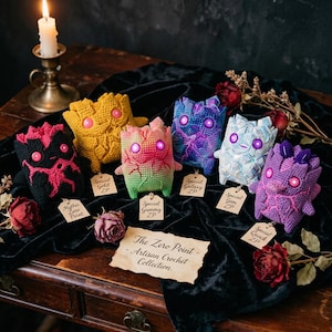May include: A collection of six small, crocheted creatures with glowing pink eyes, each featuring a unique color pattern including black, yellow, pink-green, blue-purple, white, and purple. Each creature has a small paper tag with text.