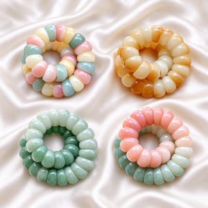 May include: Four spiral hair ties arranged in a square formation. The top left tie features a mix of pastel pink, blue, and cream segments. The top right tie is amber and cream. The bottom left is shades of green. The bottom right is pink and green.