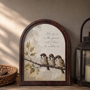 May include: An arched wooden frame containing a painting of three brown and white sparrows perched on a branch. The artwork features the text, His eye is on the sparrow, and I know he watches me, set against a textured, neutral-toned background.