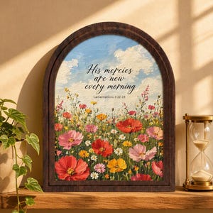 May include: An arched wooden frame containing an oil painting of a vibrant wildflower meadow under a blue sky with white clouds. The text "His mercies are new every morning" and "Lamentations 3:22-23" is printed in black in the center of the painting.