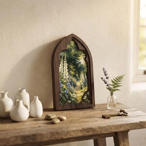 May include: A dark wood gothic arch frame containing a painting of a lush forest path with white foxglove flowers, blue blossoms, and green foliage. The frame is displayed alongside three white ceramic vases, a small glass vase with lavender and fern.