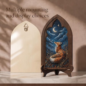 May include: An arched wooden frame containing a painting of a fox looking up at a crescent moon in a dark blue cloudy sky. The frame has a dark wood finish and sits on a matching stand. A light wood back panel with a metal hanger is also shown.