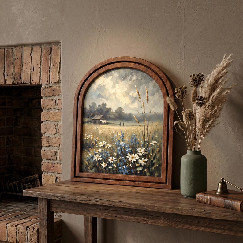 May include: An arched wooden framed painting depicting a landscape with a field of white and blue wildflowers, tall grasses, and a distant house under a cloudy sky. The artwork is displayed alongside a green vase filled with dried pampas grass.