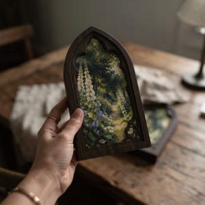 May include: A dark wood frame with a pointed gothic arch top containing a printed image of a lush garden path. The scene features tall white foxglove flowers, green foliage, and blue blossoms along a narrow dirt trail leading into the woods.