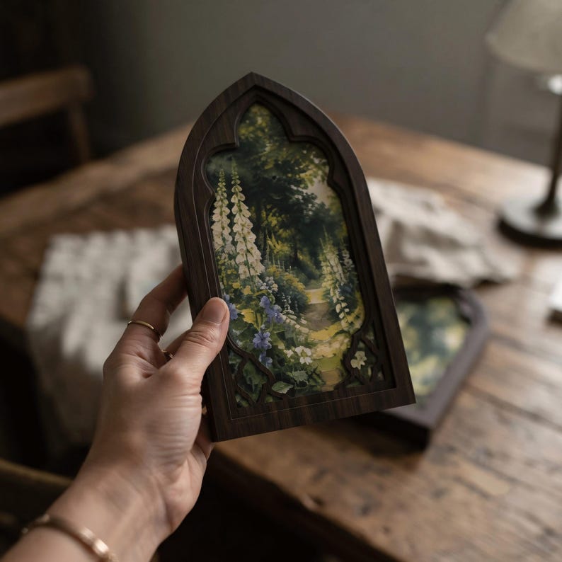 May include: A dark wood frame with a pointed gothic arch top containing a printed image of a lush garden path. The scene features tall white foxglove flowers, green foliage, and blue blossoms along a narrow dirt trail leading into the woods.