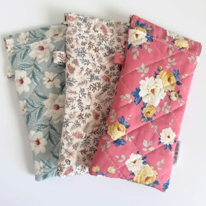 May include: Three rectangular quilted fabric pouches for glasses. The left pouch is light blue with white flowers. The center pouch features a dense pattern of small brown and grey leaves. The right pouch is pink with yellow and white floral prints.